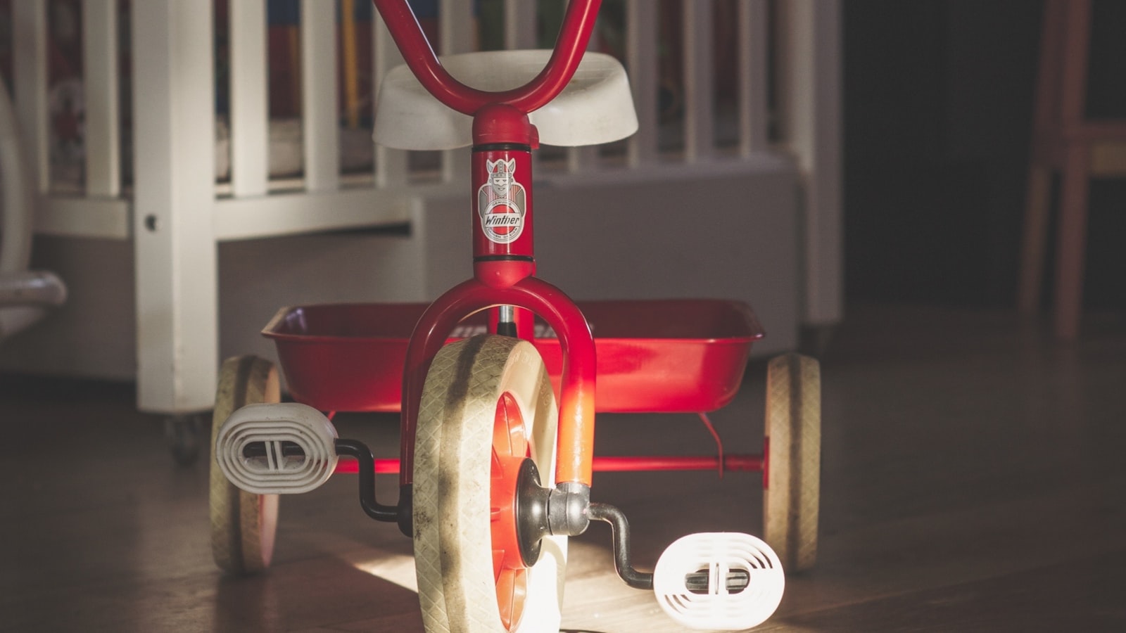 Vintage red Winther tricycle on hardwood floor with a white crib in the background — the kind of indoor scene where lead exposure most often happens in pre-1978 housing.